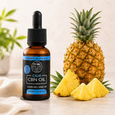An amber glass dropper bottle of NUR LIFE CBD CALM CBN OIL in Pineapple Whisk Flavor, styled next to a whole pineapple and three pineapple slices on a light beige surface with a soft neutral background.
